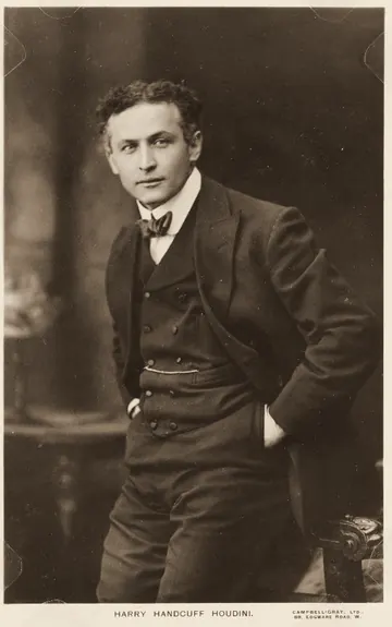 Houdini in a dark suit and bow tie, leaning on a chair, about 1913.