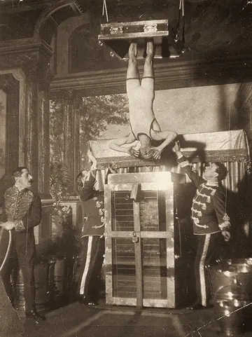Houdini hangs upside down by his feet over the Water Torture Cell while three men in uniform watch.